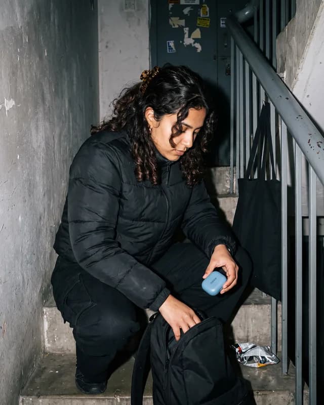 A young woman crouched in a grey stairwell, the sky blue earbuds case in her hand
